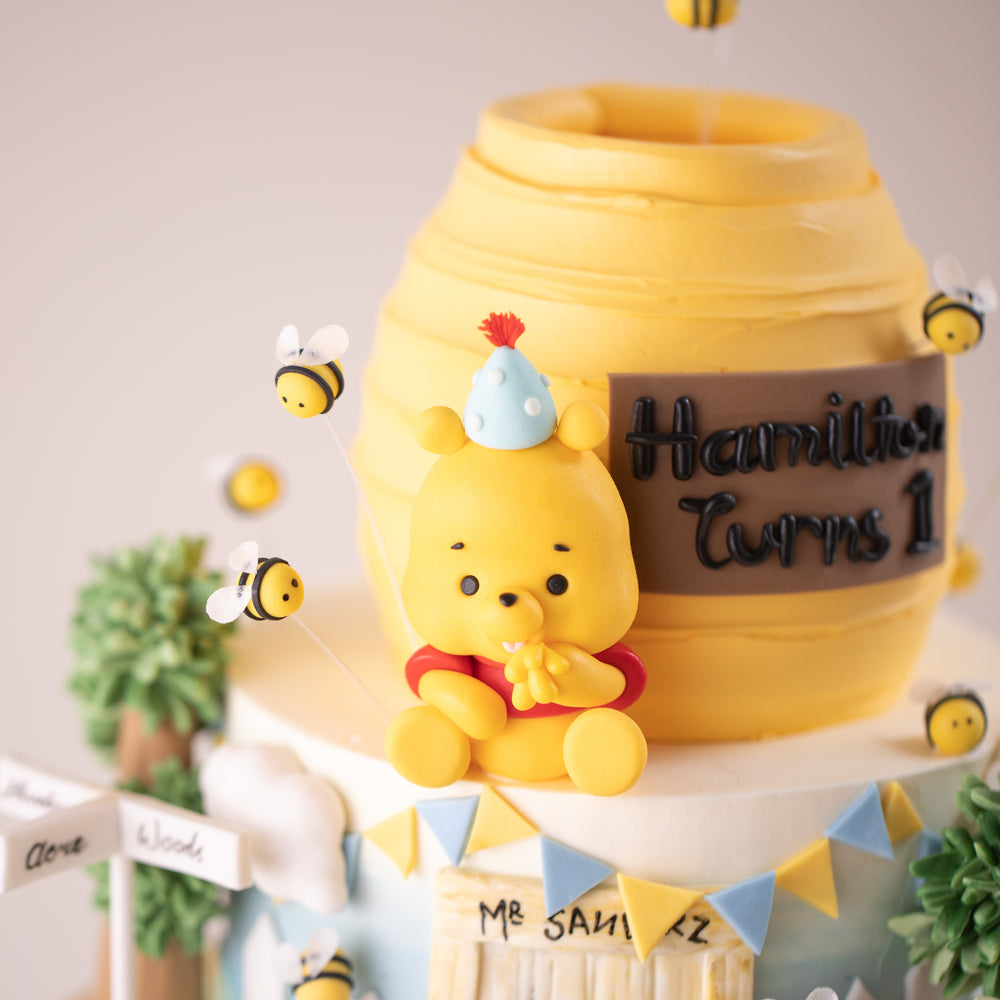 
                  
                    Load image into Gallery viewer, A fondant Winnie the Pooh eating some honey from mis paws. He is wearing a little blue party hat. There are many fondant bees flying around him cutely.
                  
                
