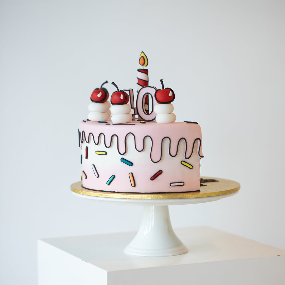A light pink cake decorated to look like pop art, with black outlines made with fondant. The cake has rainbow sprinkles, cream and cherries, all done with fondant. There is a single red and white striped candle in the middle that is also made with fondant.