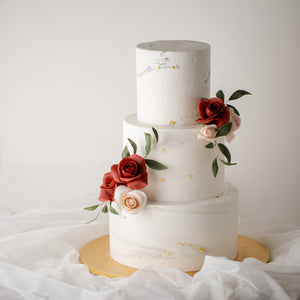 
                  
                    Load image into Gallery viewer, A tall three tier minimalistic wedding cake, with a white and light grey marble buttercream base. The cake has some bright red and light pink edible sugar roses, along with some darker green edible sugar leaves. The white and grey marbled buttercream has some edible gold leaf carefully placed, to give it a stunning contrast. 
                  
                