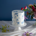 A cake with a white fondant base and light blue handpainted details. The cake decorations resemble a royal copenhagen teapot, and it looks like fine china.