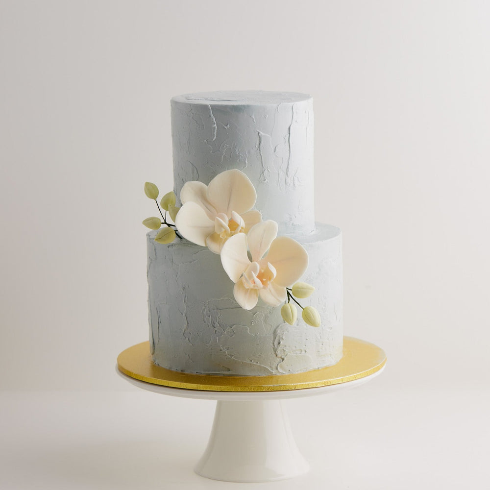 
                  
                    Load image into Gallery viewer, A 2 tier wedding cake with a light cloudy grey buttercream base that has texture to it. The cake has two light peach edible sugar orchids in the middle, along with some pale green edible orchid buds. The orchids look incredibly realistic.
                  
                