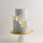 A 2 tier wedding cake with a light cloudy grey buttercream base that has texture to it. The cake has two light peach edible sugar orchids in the middle, along with some pale green edible orchid buds. The orchids look incredibly realistic.