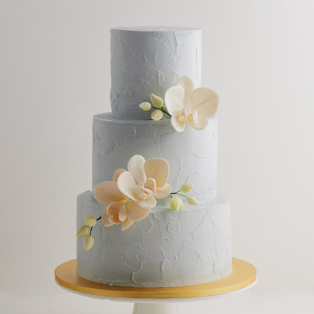 A 3 tier wedding cake with a light cloudy grey buttercream base that has texture to it. The cake has three light peach edible sugar orchids in the middle, along with some pale green edible orchid buds. The orchids look incredibly realistic.