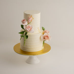 
                  
                    Load image into Gallery viewer, A two tier white buttercream wedding cake. The buttercream has white horizontal lines to give it texture. There are light pink edible sugar roses with green stems and leaves, and they look incredibly realistic.
                  
                