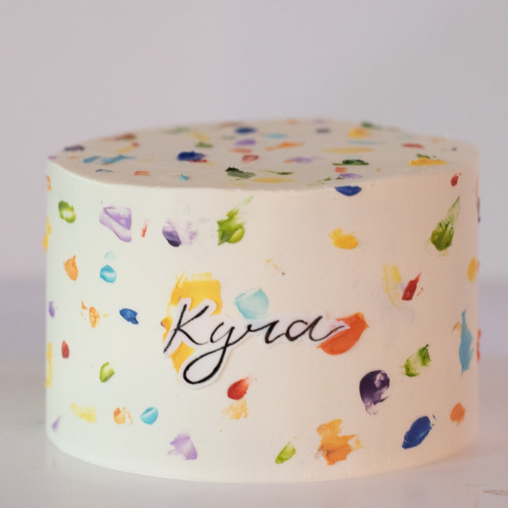A white buttercream cake with rainbow buttercream dots painted all around the cake, terrazzo style. The rainbow dots resemble paint splotches onna canvas. In the middle of the cake, there is a name 