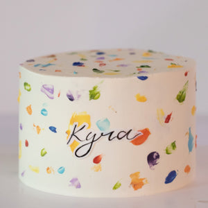 
                  
                    Load image into Gallery viewer, A white buttercream cake with rainbow buttercream dots painted all around the cake, terrazzo style. The rainbow dots resemble paint splotches onna canvas. In the middle of the cake, there is a name &amp;quot;Kyra&amp;quot; written on edible icing sheet.
                  
                