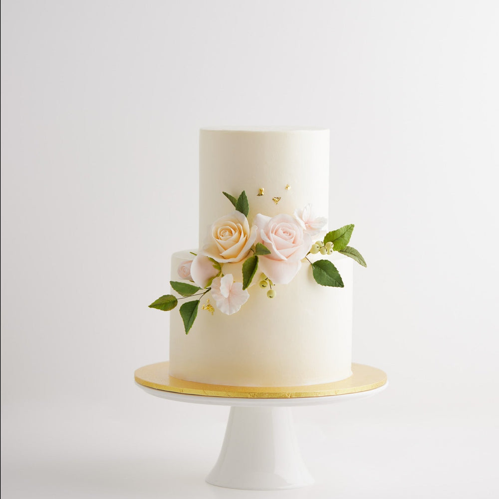 A two tier wedding cake with smooth white buttercream coating. There are light pink and blush edible sugar roses on the cake, along with green edible sugar leaves. The roses are incredibly realistic.