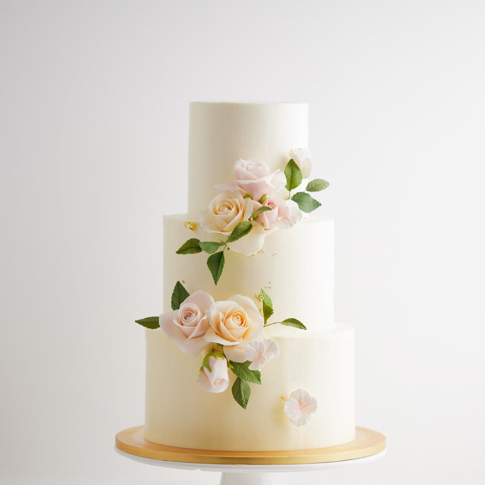 A three tier wedding cake with smooth white buttercream coating. There are light pink and blush edible sugar roses on the cake, along with green edible sugar leaves. The roses are incredibly realistic.