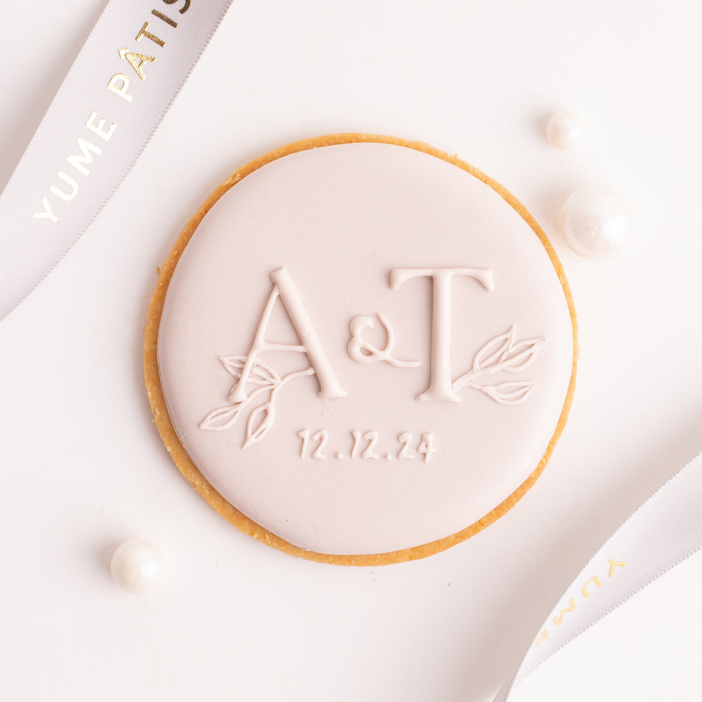 A round cookie with the initials "A&T" on it in cursive. Below the initials is the date "12.12.24". The cookie is decorated with royal icing in a soft, pastel pink.