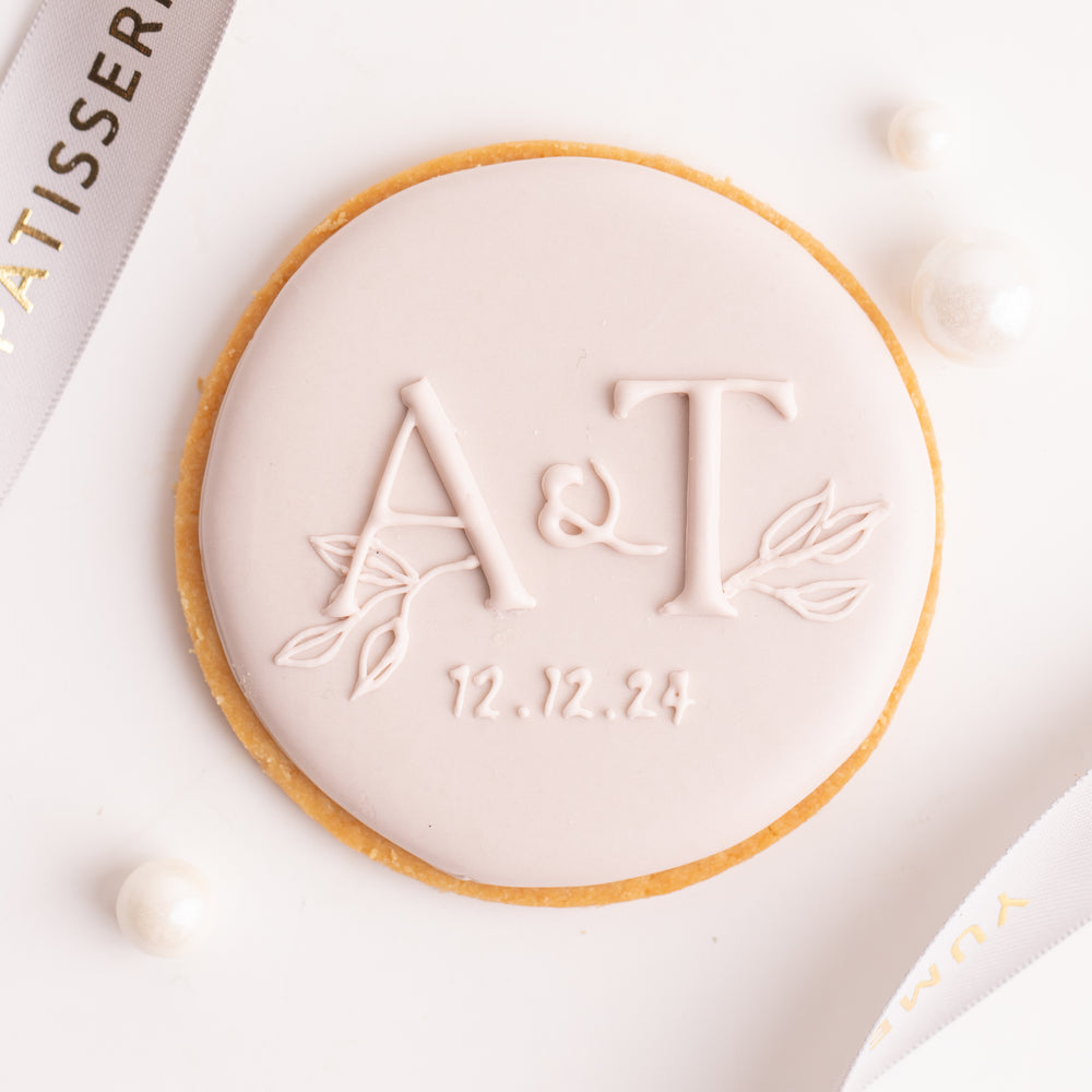 A round cookie with the initials "A&T" on it in cursive. Below the initials is the date "12.12.24". The cookie is decorated with royal icing in a soft, pastel pink.