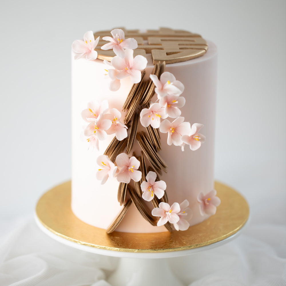 A slim and tall cake with a light pink fondant base. The cake has gold scallops climbing up the side of the cake, along with some light pink sugar plum blossoms. The top of the cake has 