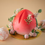 A cake carved into the shape of a peach (Shou tao). The cake has a red to light pink gradient, and small fondant pink peaches around. It also has small light pink plum blossoms around.