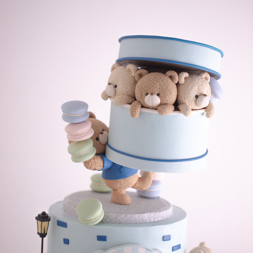 
                  
                    Load image into Gallery viewer, A close up on the top tier of the cake. It has a little fondant teddy bear carrying a large blue gift box with other smaller bears inside. The fondant teddy bear is also carrying a pile of macarons in light blue, pink and green. The fondant teddy bear is wearing a blue shirt.
                  
                