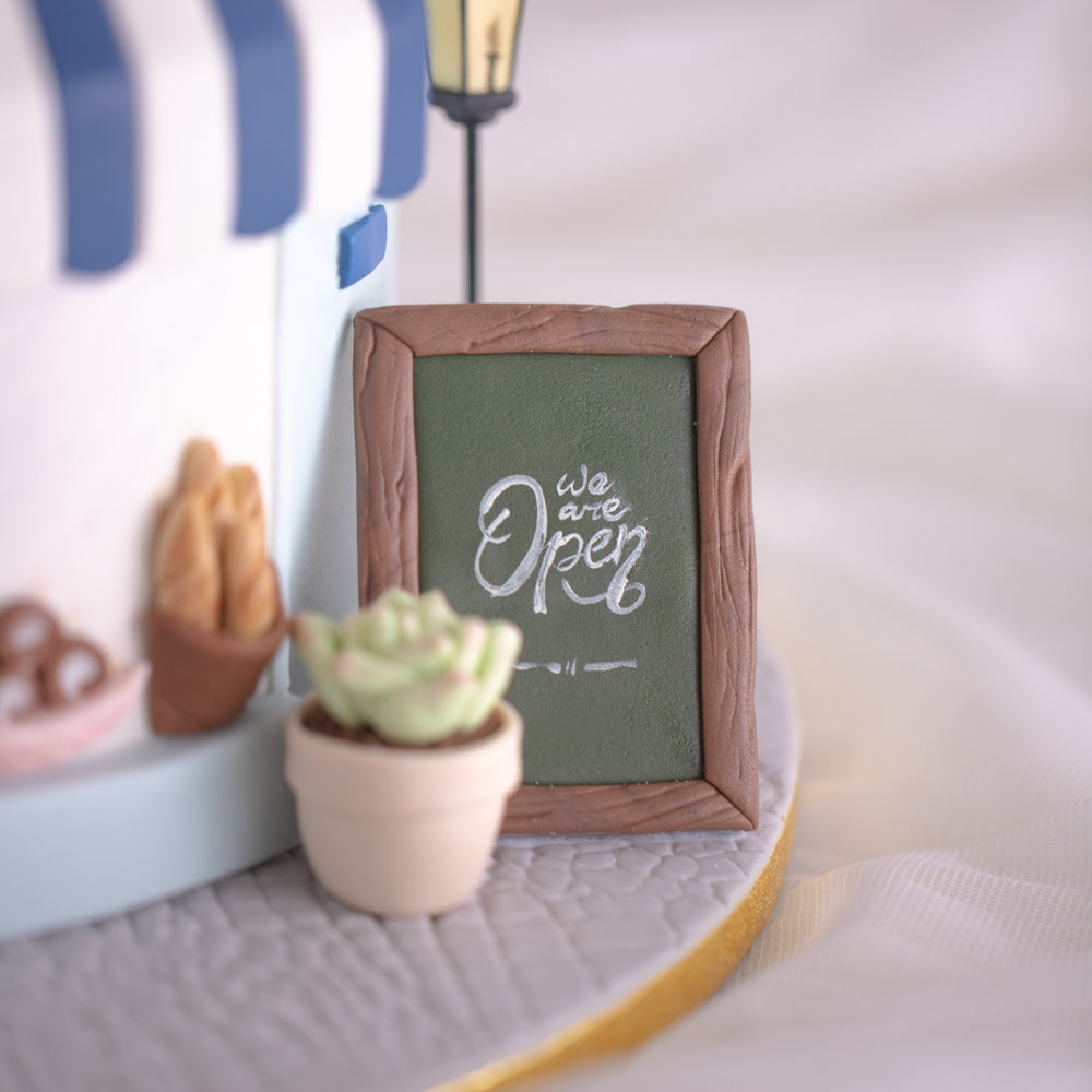 
                  
                    Load image into Gallery viewer, A close up on the fondant cafe signboard on the bottom of the cake. It has the words &amp;quot;We are Open&amp;quot; hadnpainted on it that resemble chalk on a chalk signboard. In front of the signboard there is a tiny fondant potted plant, along with some fondant breads on a windowsill.
                  
                