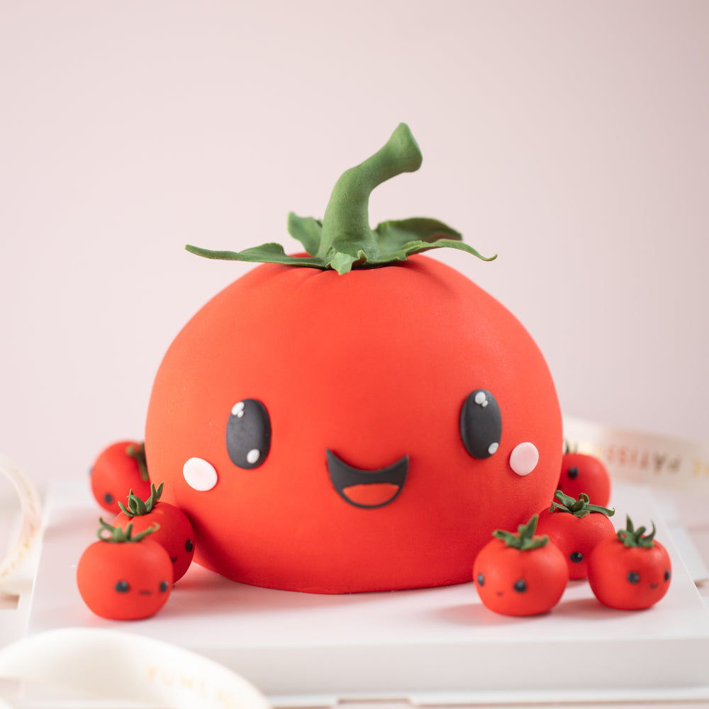 A cake that has been carved and decorated to look like a tomato. It has an adorable smiling face in the middle, and pink blushing cheeks. There are many smaller cherry tomatoes scattered around the cake board, each of them having different facial expressions.